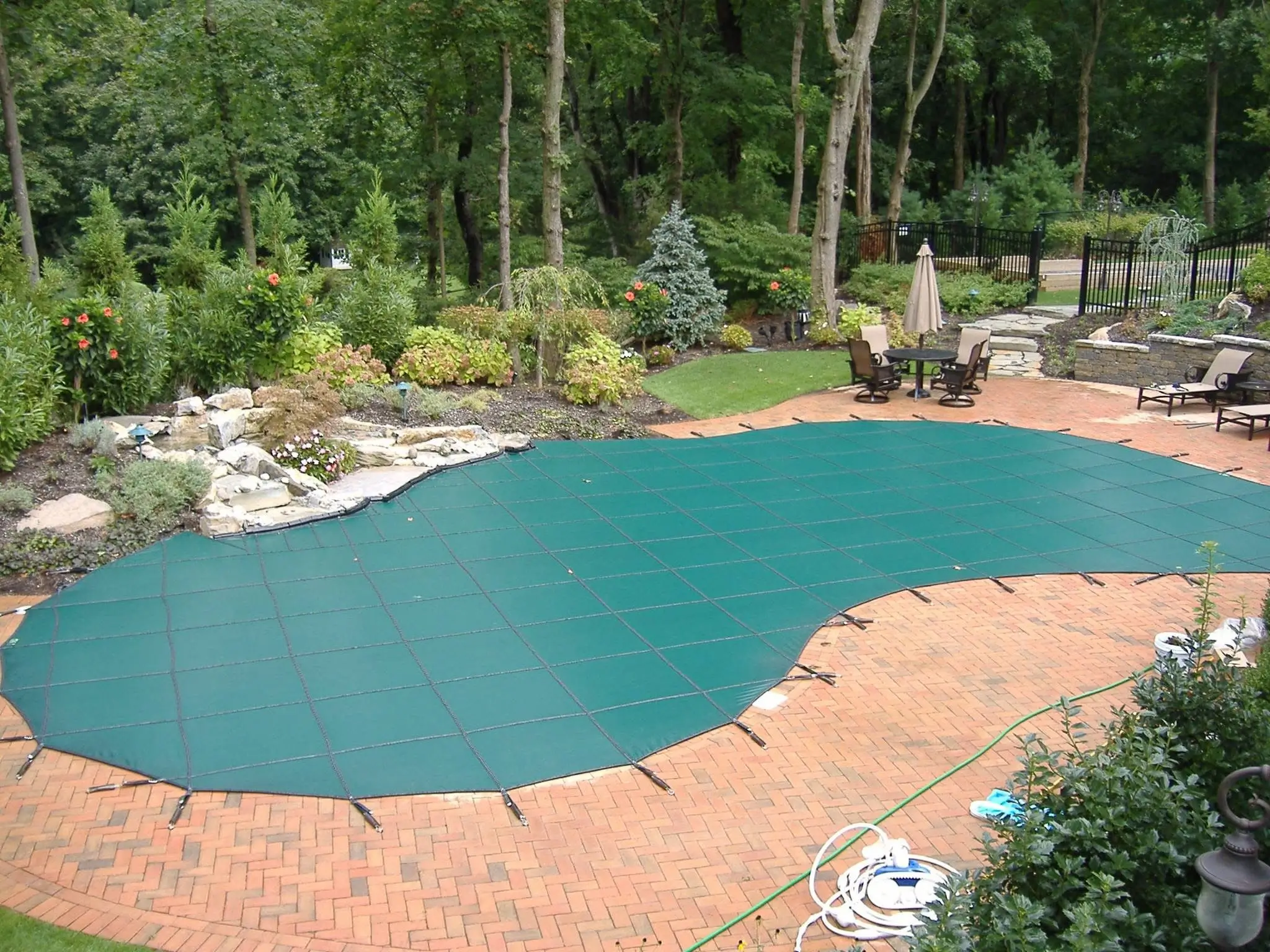 Green mesh safety cover strapped over an in-ground pool next to landscaping and a seating area.