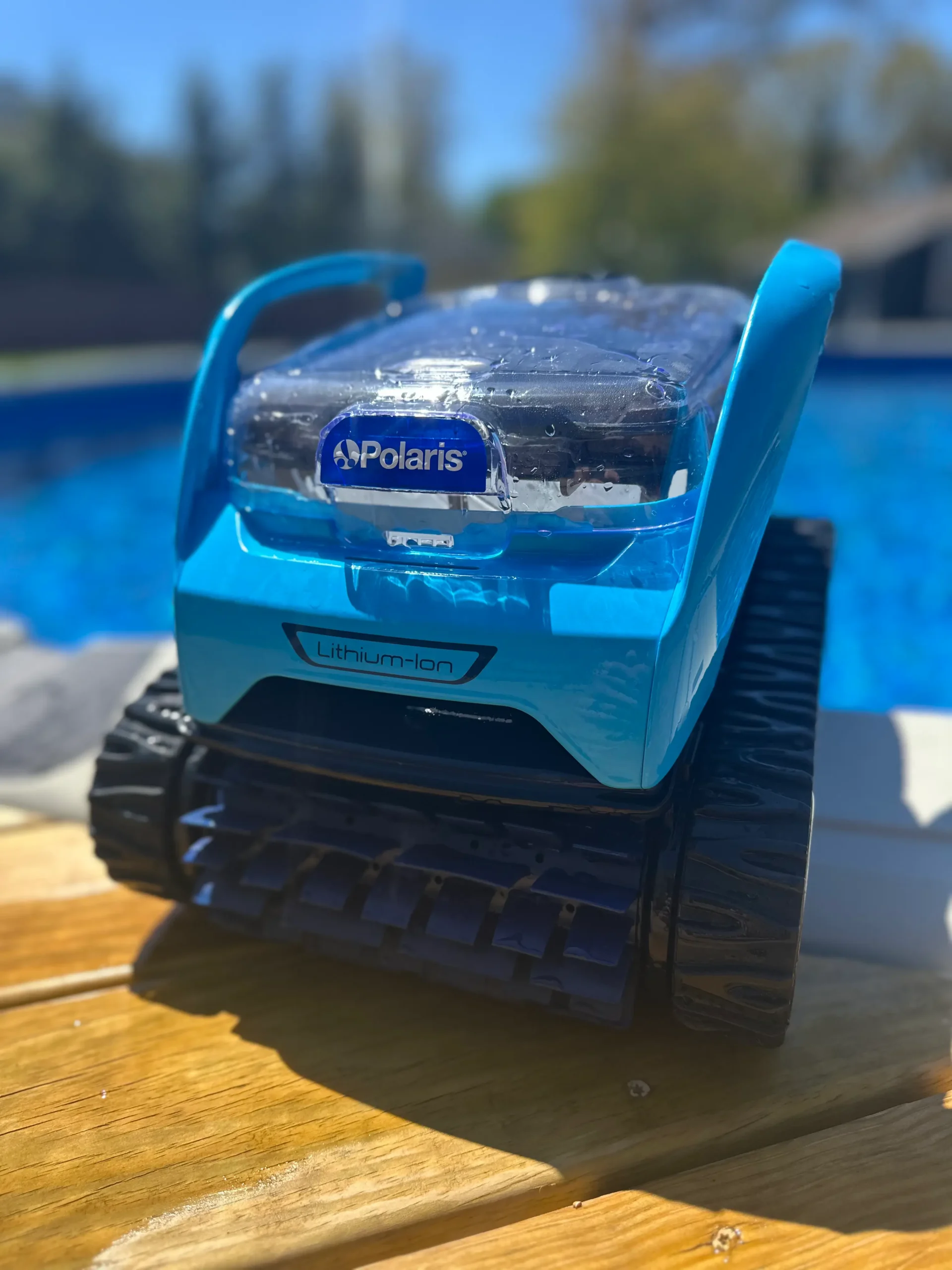 A close-up shot of a bright blue Polaris cordless robotic pool cleaner with "Lithium-Ion" printed on the front. It's resting on a light brown wooden deck with a swimming pool in the background.