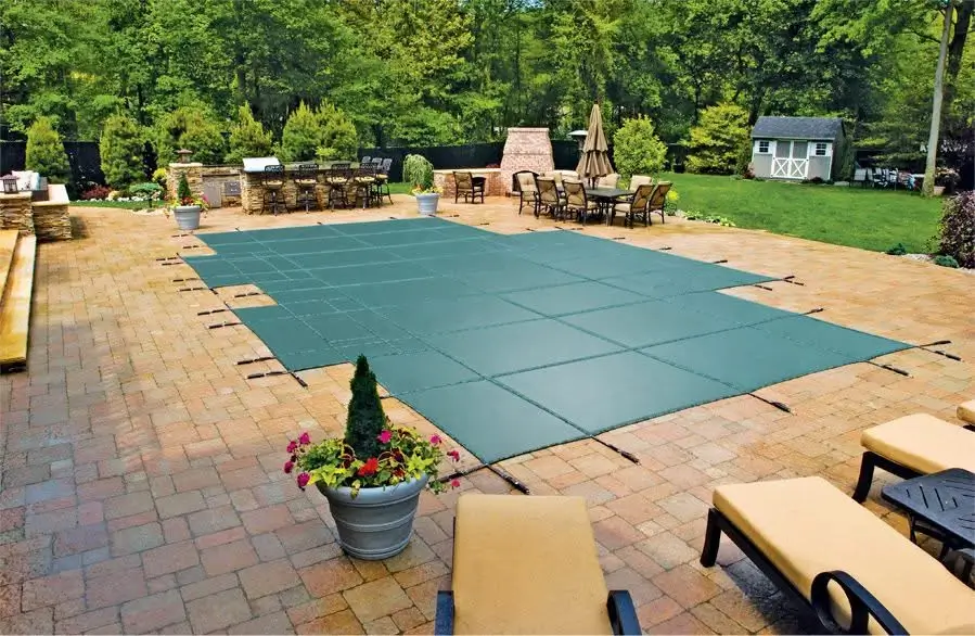 Green mesh pool safety cover stretched across an in-ground pool on a brick paver patio with lounge chairs.